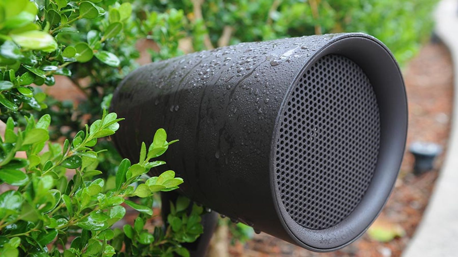A water-resistant outdoor speaker nestled among vibrant greenery, covered in water droplets.