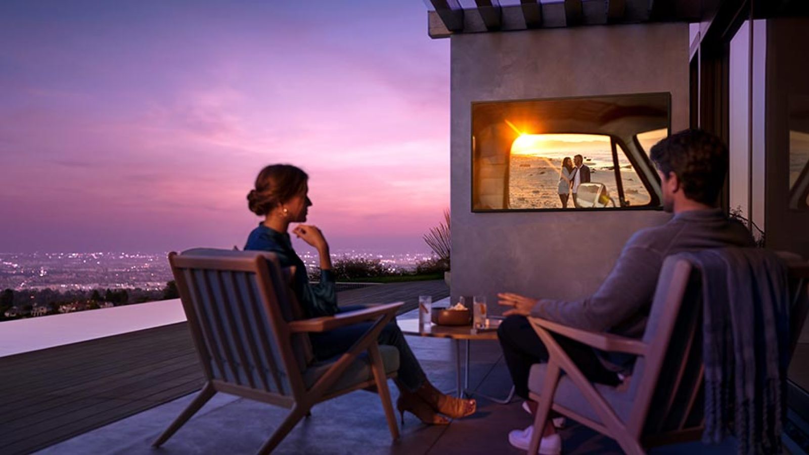A couple relaxing on a balcony at sunset, watching a romantic scene on an outdoor TV overlooking a scenic city view.