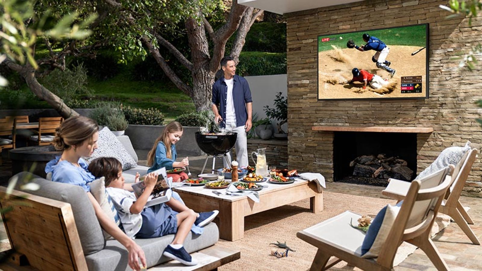 A family enjoying an outdoor living space with a grill, dining area, and a TV displaying a baseball game.