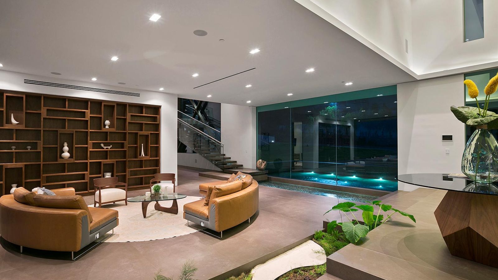 Luxurious living room with leather seating, an open bookshelf, and a view of an illuminated pool.