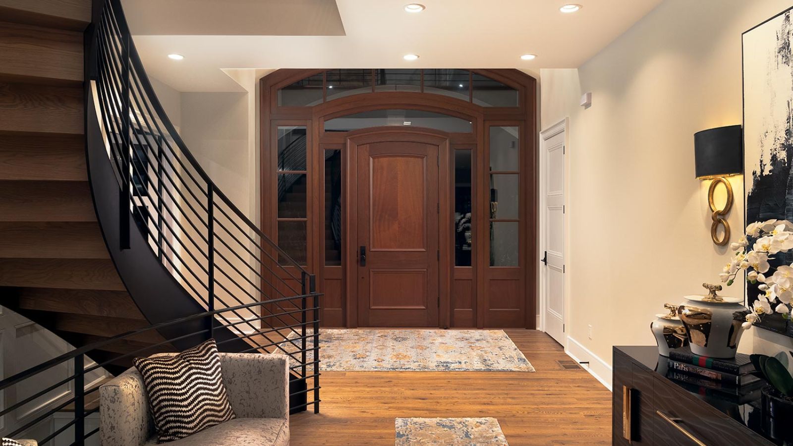 A modern entryway with a curved staircase, wooden double doors, and stylish decor under warm recessed lighting.