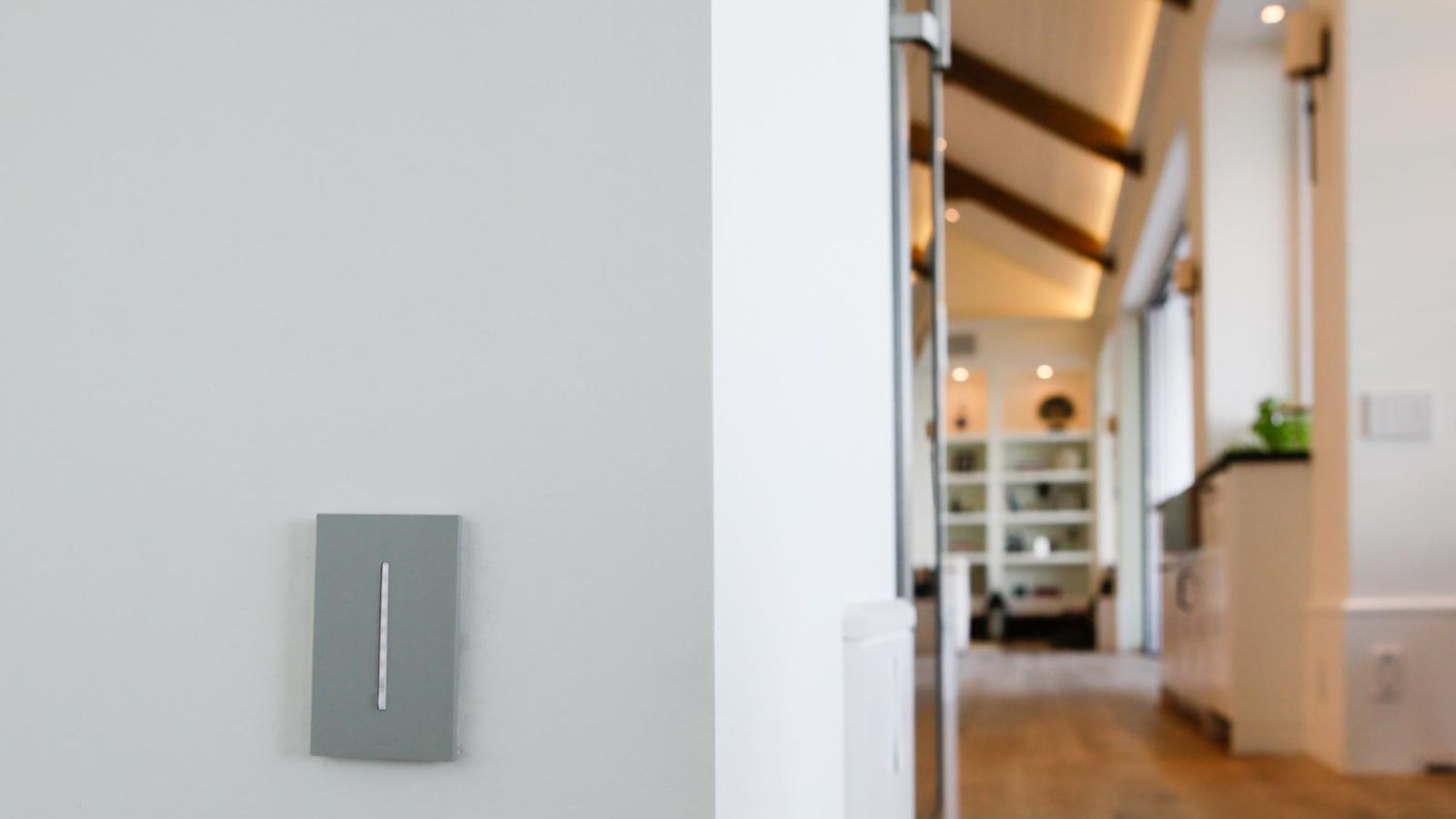 A stylish hallway with a sleek light switch on a minimalist wall, leading to a living space with exposed wooden beams and shelves.