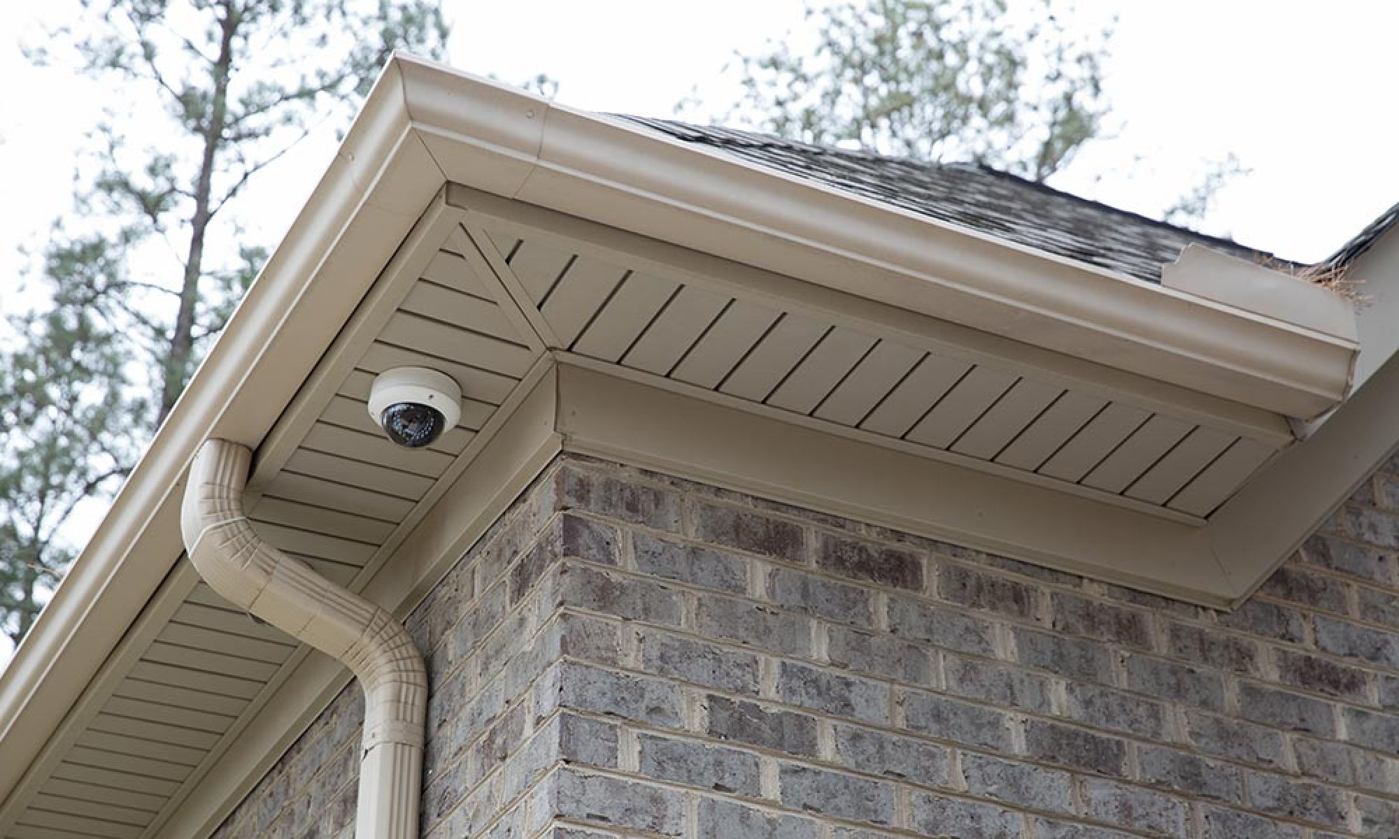 A discreet security camera installed under the eaves of a brick house exterior.