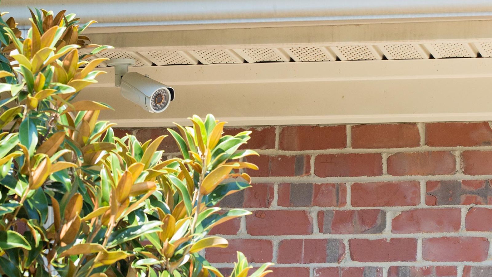 Corner-mounted security camera on the soffit of a brick home, with bushes below.