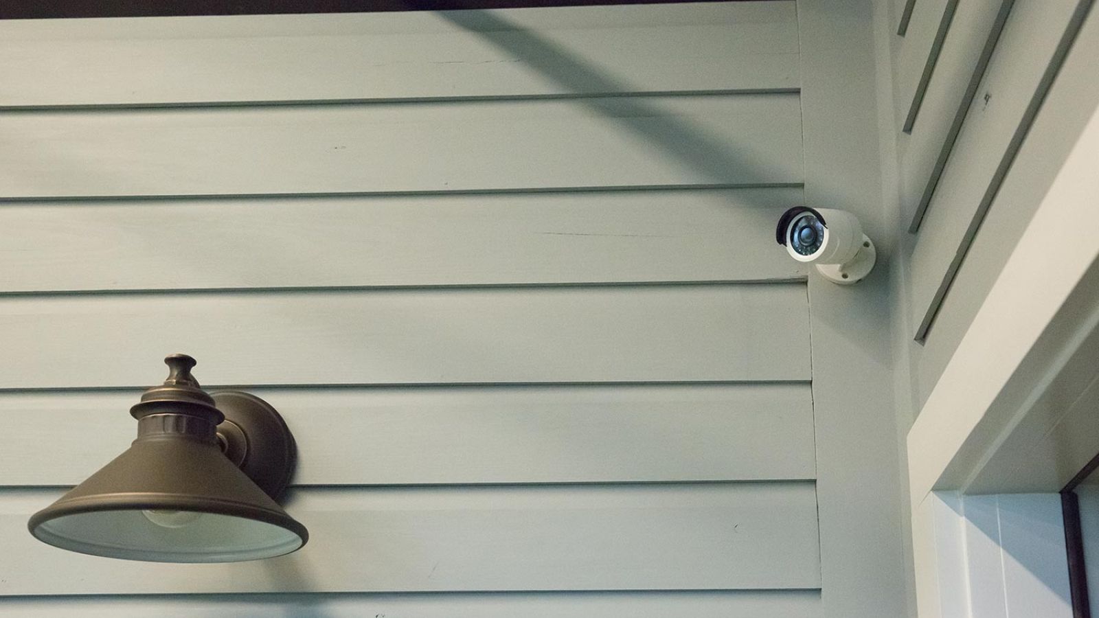 Security camera mounted on a light gray wall, next to a classic outdoor wall light fixture.