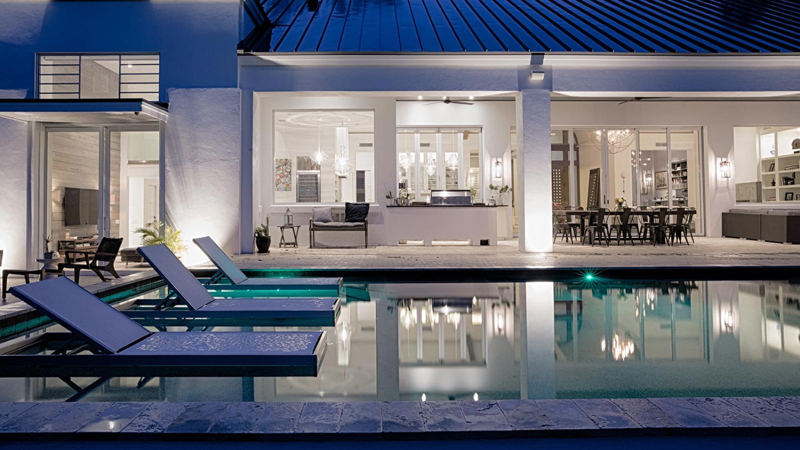 An elegant outdoor pool area at night, with illuminated interiors showcasing modern design and ample seating.