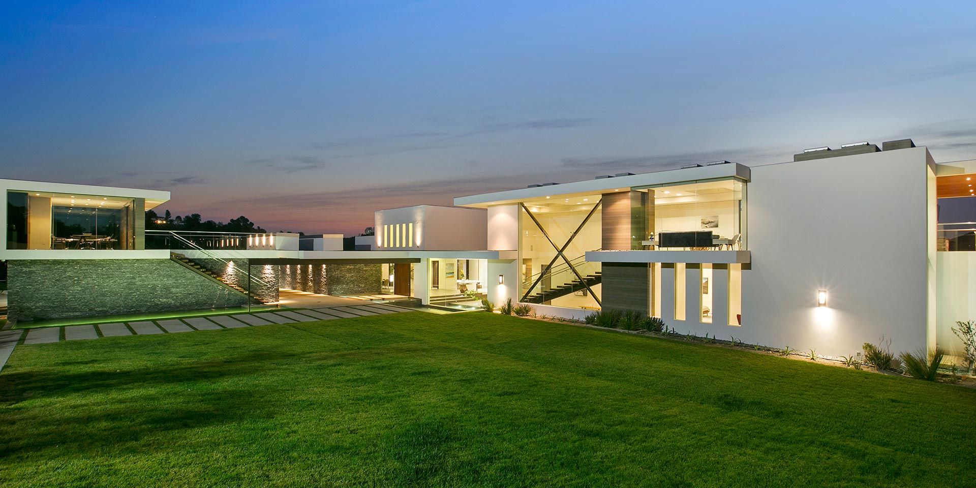 A large, modern home illuminated at dusk, showcasing expansive glass windows and open architectural design.