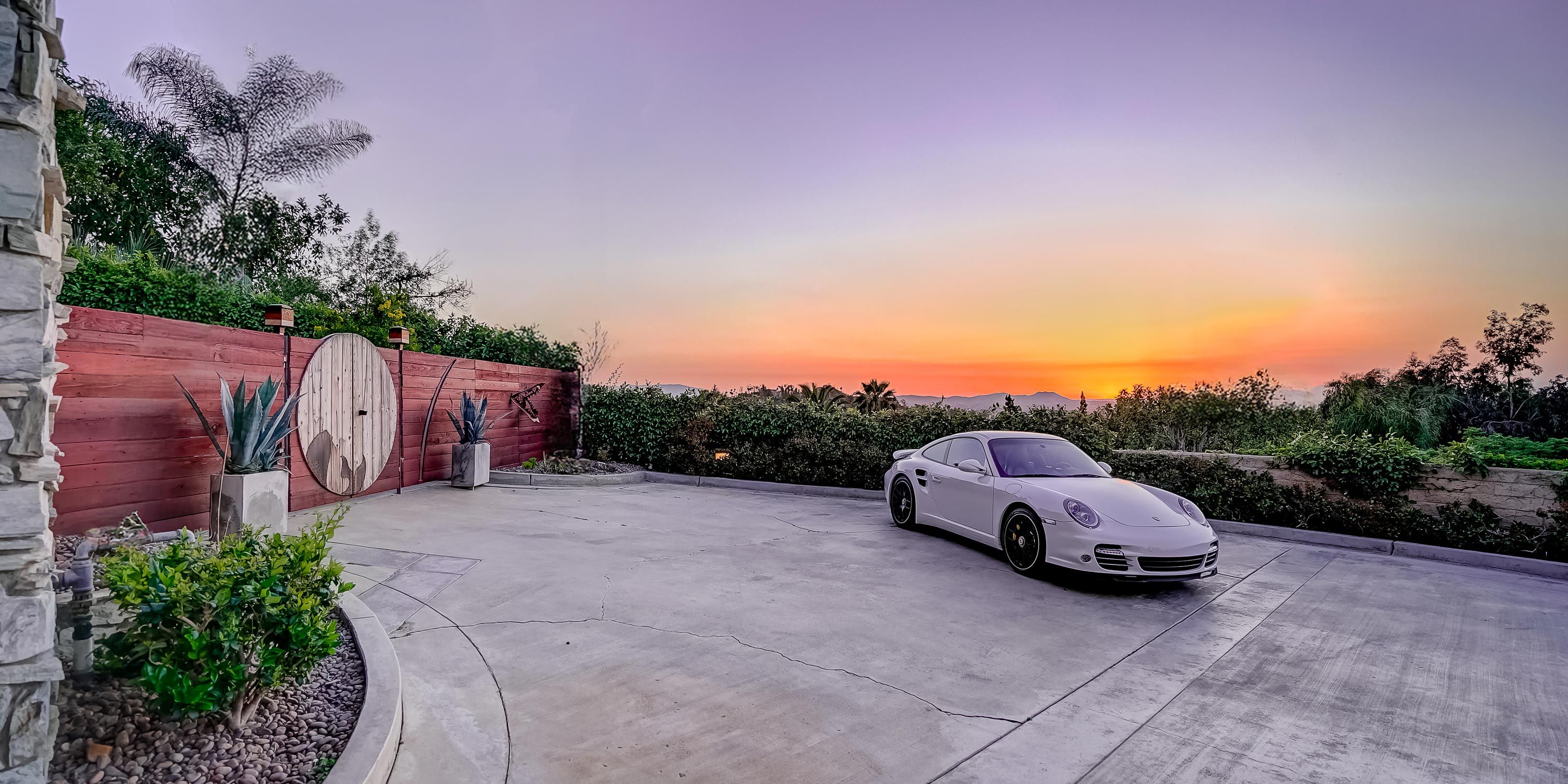 A spacious driveway with a luxury car parked, framed by a wooden gate and plants, with a sunset backdrop.