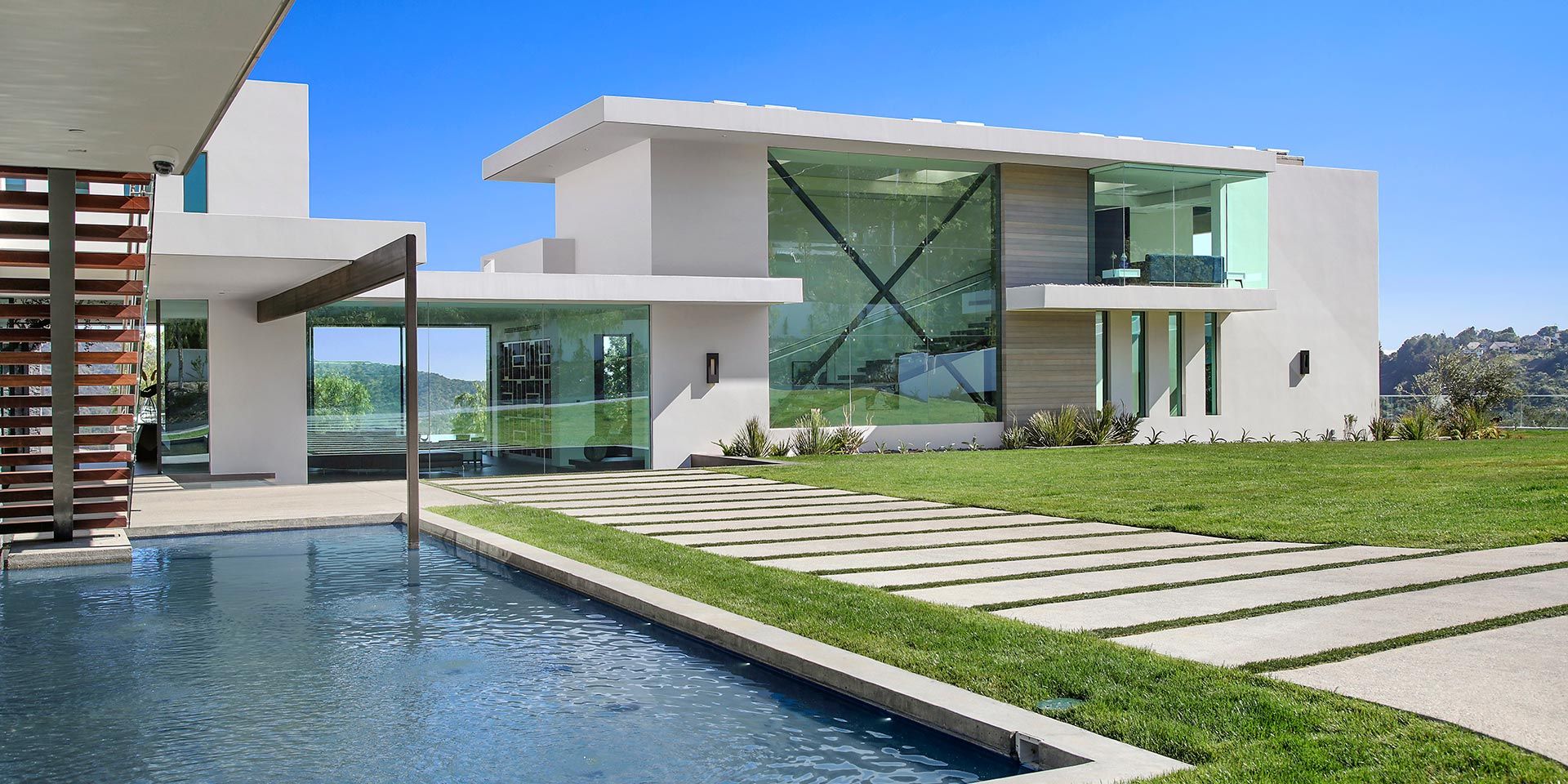 A modern house with large glass windows, a well-manicured lawn, and a pool running alongside the walkway.