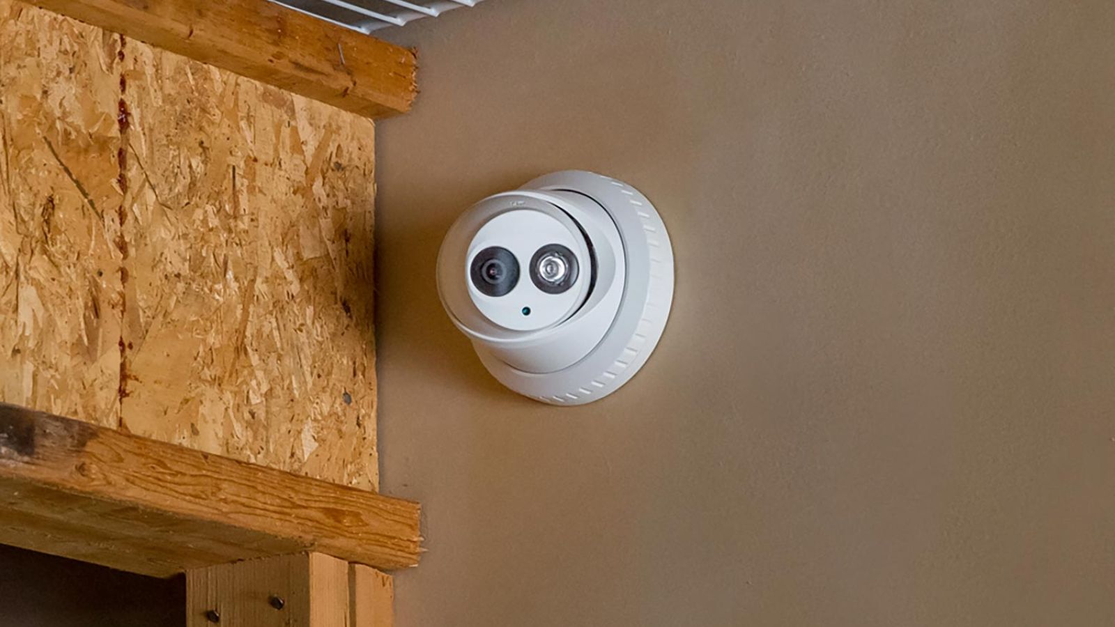 Indoor dome camera mounted on a beige wall, near a wooden beam and wire shelving.