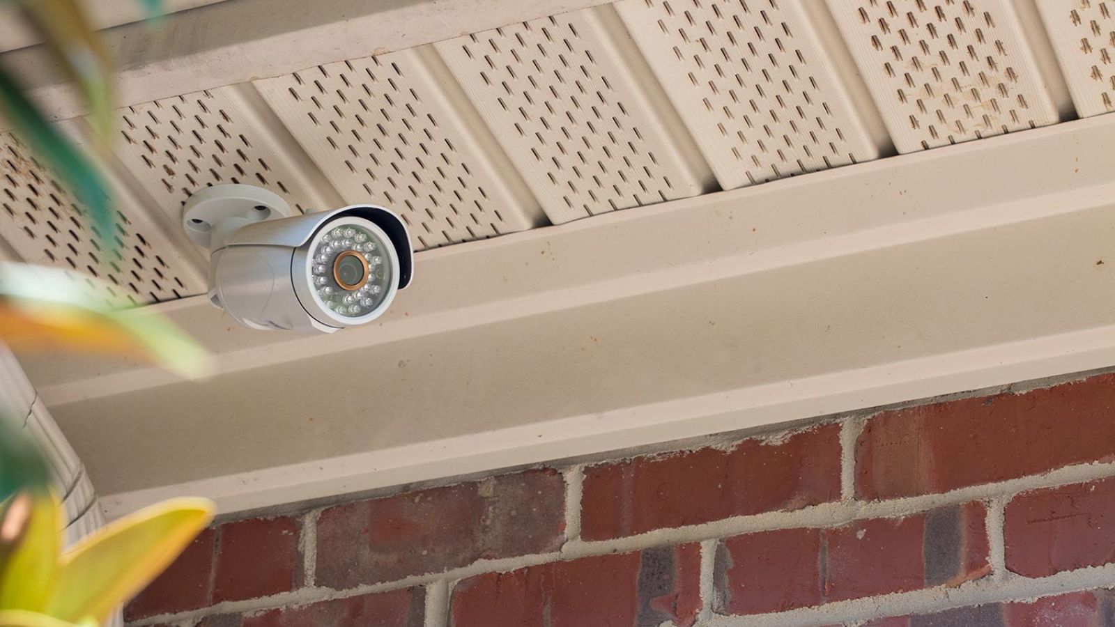 Corner-mounted security camera on the soffit of a brick home, with bushes below.