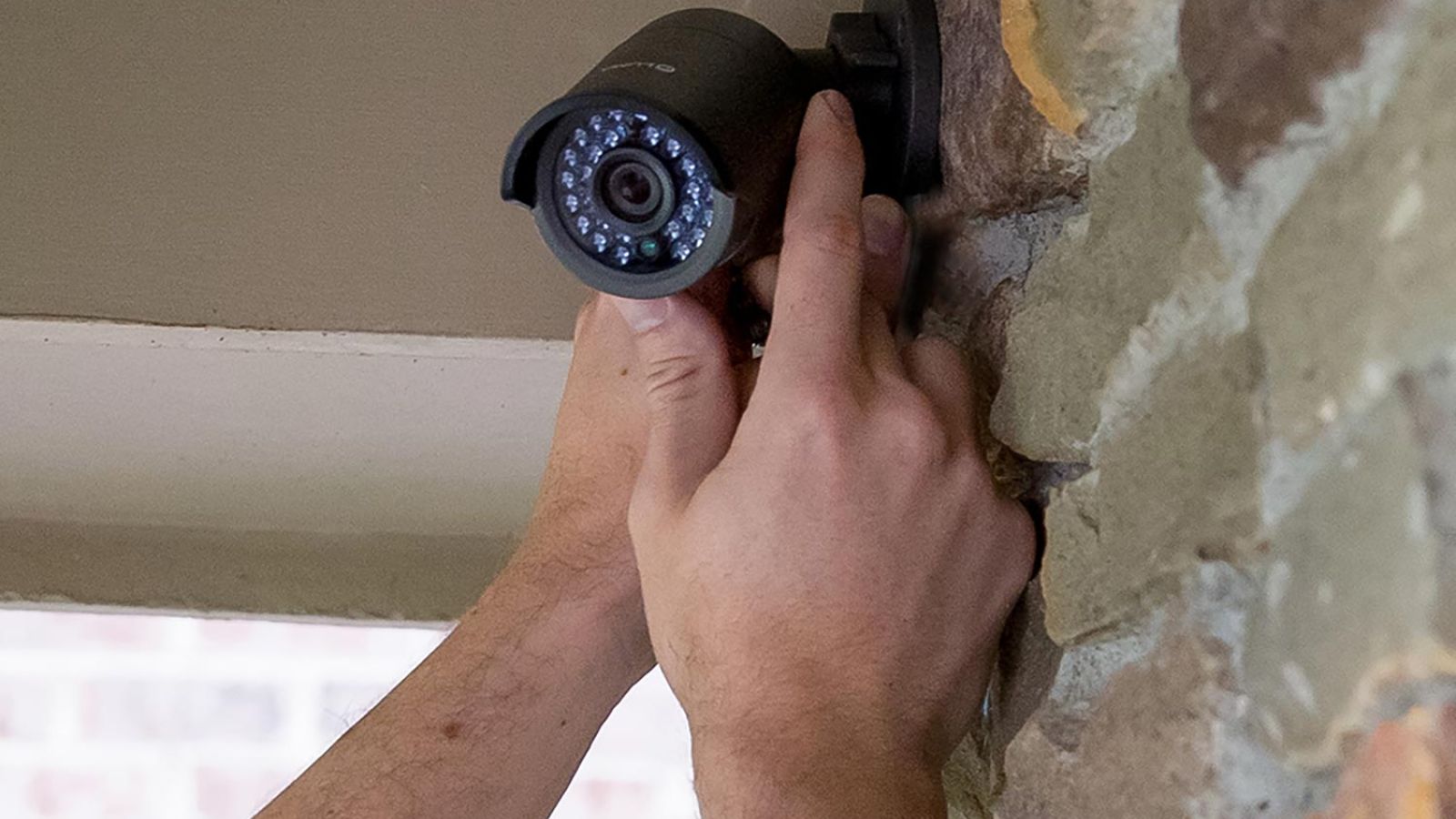 Hands installing a black outdoor security camera on a stone wall.