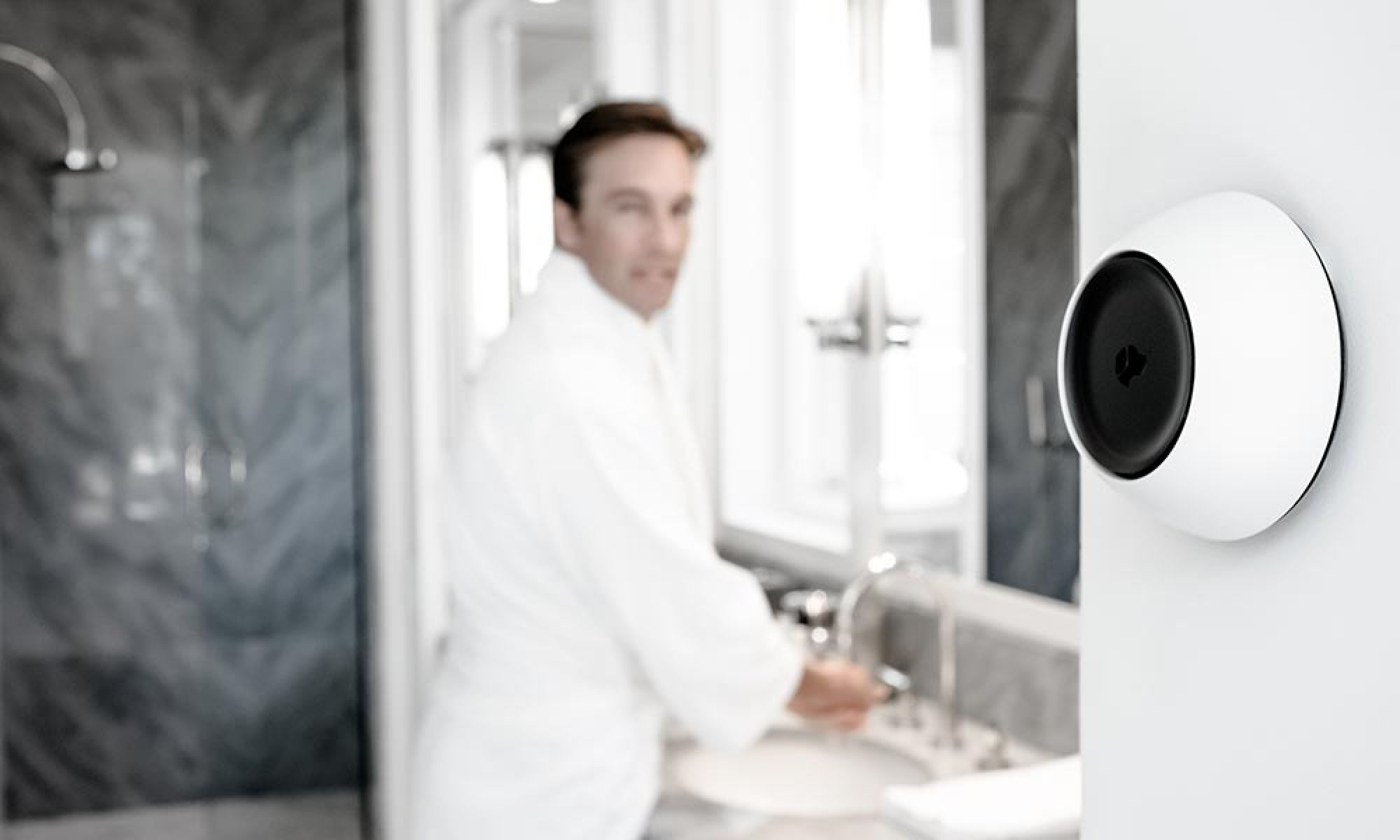 A bathroom scene with a futuristic wall-mounted device in focus, and a person in a bathrobe washing hands in the background.