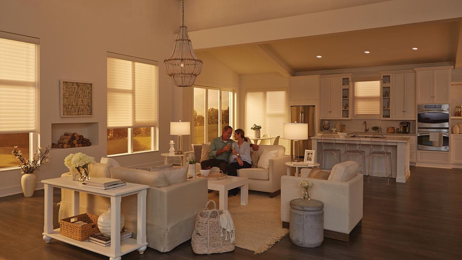 A bright living room with white furniture, illuminated by warm light through window shades, overlooking a serene countryside.