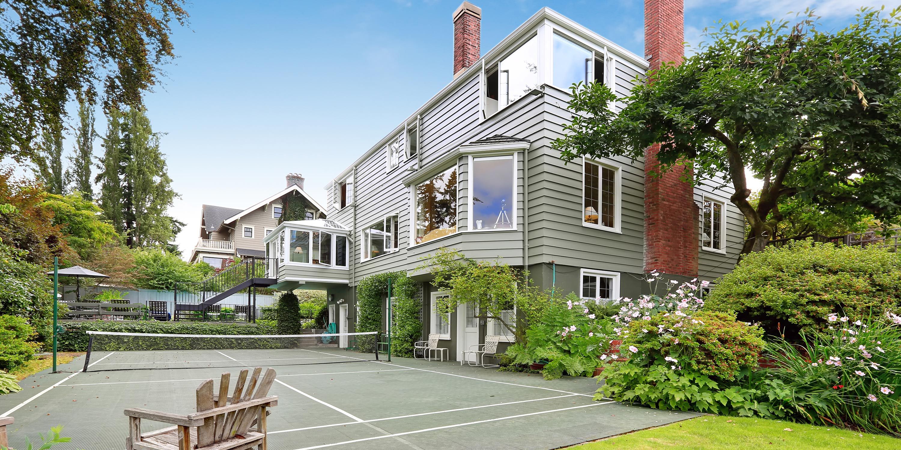 A large, well-maintained tennis court in the backyard of a multi-story house surrounded by greenery.